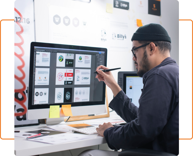 A man sitting at a desk, focused on a computer screen, holding a pen in his hand, Working on a graphic design service project.